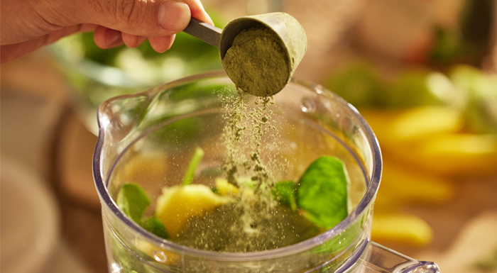 Scoop of green powder being added to a blender of fruit and spinach