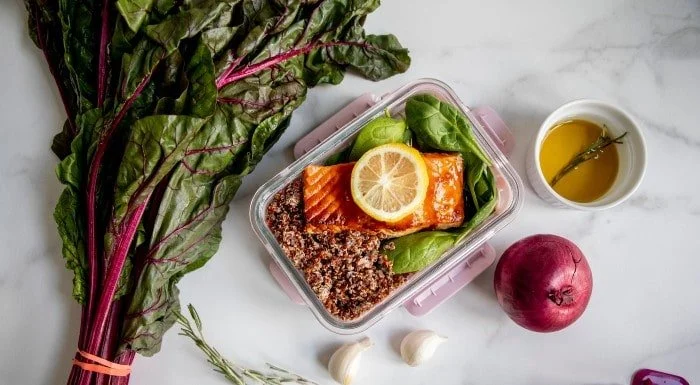 Meal prep container of salmon, quinoa and greens, next to Swiss chard and a red onion