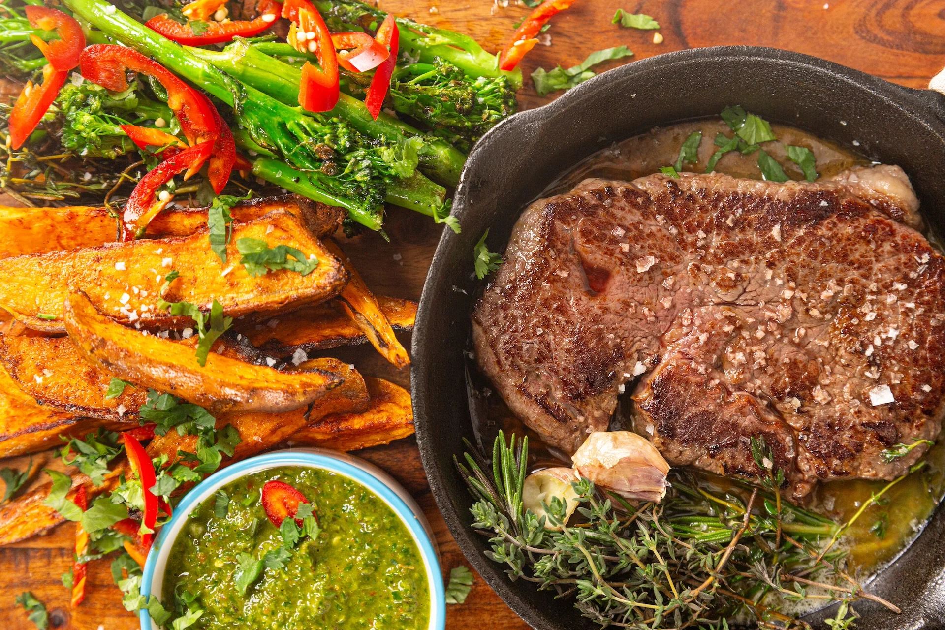 Cooked steak in a pan with sweet potatoes fries, broccolini and chimichurri sauce.